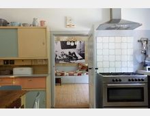 Küche im Retro-Stil mit pastellfarbenen Schränken, einem Gasherd mit Metallabzug und gefliester Rückwand. Im Hintergrund ein angrenzender Raum mit Holzboden, einem Tagesbett mit rot-weiß karierter Decke und einem Schwarz-Weiß-Bild an der Wand.