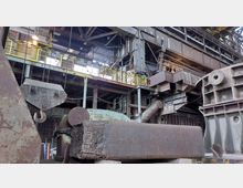 Industriehalle mit rostigen Metallstrukturen, großen Maschinen und einem schweren Hakenkran im Vordergrund. Im Hintergrund sind Geländer, Stahlträger und Rohre zu sehen, die typisch für eine alte Fabrikumgebung sind.