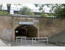 Backsteintunnel unter einer Bahnbrücke mit einem Schild "Berlin-Grunewald" und Verbotsschildern darüber; der Tunnel führt zu einem S-Bahnhof, umgeben von Bäumen und Geländern.