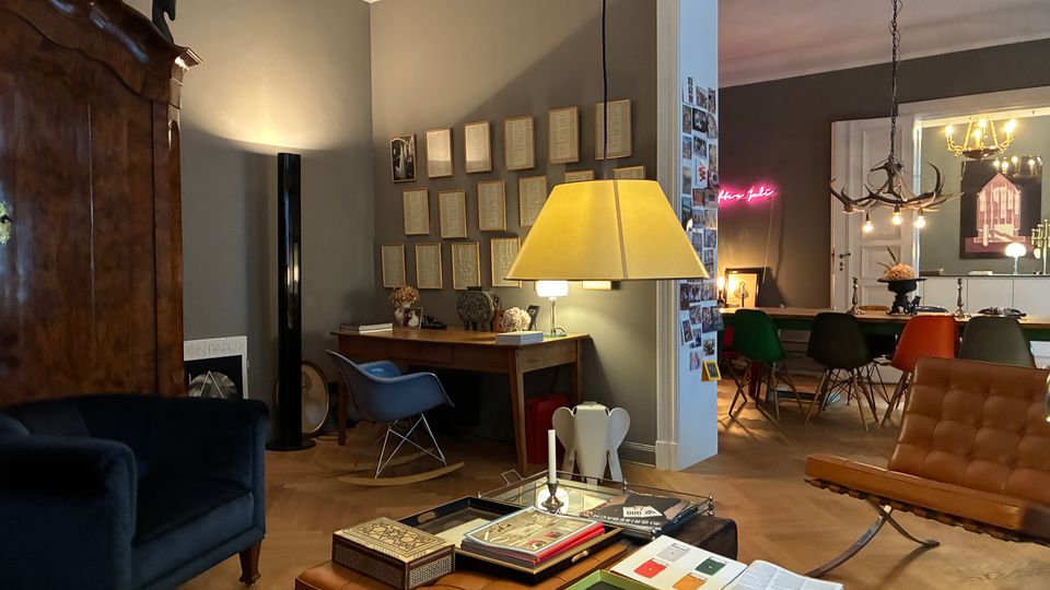 Wohnzimmer mit grauen Wänden, Stuckdecke und Holzboden, eingerichtet mit großem Holzschrank, Sesseln, Lederhockern und Couchtisch unter einer großen Hängelampe. Im Hintergrund führt eine breite Türöffnung in ein Esszimmer mit langem Tisch, bunten Stühlen und einem Geweih-Kronleuchter.