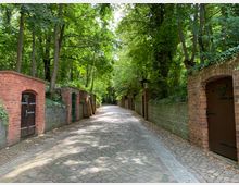 Gepflasterter Weg durch eine von Bäumen gesäumte Allee mit roten Ziegelmauern und Holztüren auf beiden Seiten. Die Umgebung ist von dichtem Grün und Schatten geprägt.