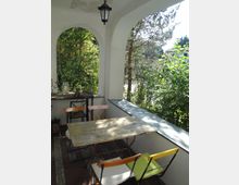 Überdachte Veranda mit weißen Rundbögen und hängender Laterne, ausgestattet mit einem kleinen Holztisch und Stühlen auf Fliesenboden. Seitlich öffnet sich der Blick ins dichte Grün von Bäumen und Sträuchern, mit einem niedrigen Geländer entlang der Kante.