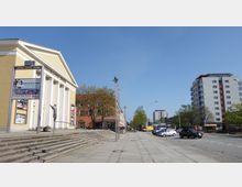 Gelb gestrichenes Gebäude mit Säulenfront und der Aufschrift „Friedrich-Wolf-Theater“ auf der linken Seite, umgeben von weiteren niedrigen Gebäuden und Bäumen. Rechts im Bild sind Wohngebäude und parkende Autos entlang einer Straße zu sehen, mit klarem Himmel im Hintergrund.