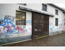 Weiße Hausfassade mit mehreren Graffitis, großen Holztoren in der Mitte und vergitterten Fenstern. Der Boden davor ist mit Kopfsteinpflaster bedeckt.