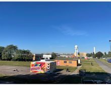 Weitläufige Parklandschaft mit vereinzelten Gebäuden, darunter ein bunt bemalter Container mit der Aufschrift „Wir machen was“, und im Hintergrund ein Turm mit einer kugelförmigen Spitze sowie ein hoher, schmaler Betonbau. Der Himmel ist klar und blau.