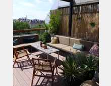 Dachterrasse mit Sitzbereich, bestehend aus einem Ecksofa mit Kissen, Holztischen und Stühlen. Pflanzen und Bambuswände rahmen den Bereich ein, mit Blick auf die umliegenden Gebäude und Bäume.