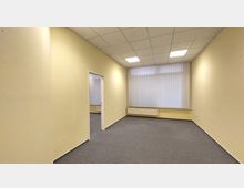 Ein leerer Büroraum mit gelb gestrichenen Wänden, grauem Teppichboden und einer Decke mit Rasterpanels und eingelassenen Leuchten. Rechts ist ein großes Fenster mit vertikalen Lamellenjalousien, darunter ein Heizkörper; eine offene Tür führt in einen weiteren Raum.