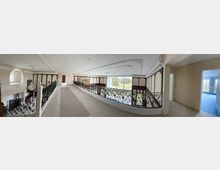 Panoramablick auf das obere Geschoss eines luxuriösen Gebäudes mit verzierten Geländern aus schwarzem Metall, einem Kronleuchter und eleganten Wandpaneelen. Große Fenster und ein lichtdurchfluteter Raum bieten einen Blick auf eine gepflegte Rasenfläche im Außenbereich.