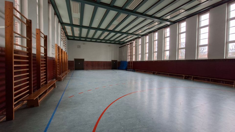 Leere Sporthalle mit großem Hallenboden und farbigen Spielfeldmarkierungen, an den Seiten mit Sprossenwänden und Bänken. Hohe Fensterreihen lassen viel Tageslicht herein, darüber eine Decke mit sichtbaren Trägern und Leuchten.
