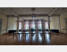 Großer, heller Raum mit verzierten Decken, säulenförmigen Ornamenten und einem auffälligen Kronleuchter. Die Fenster sind mit rosafarbenen Vorhängen dekoriert, während Tische und Stühle auf einem Holzboden arrangiert sind.