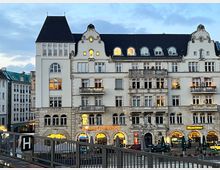 Historisches mehrstöckiges Gebäude mit verzierten Fassaden und dunklem Schieferdach, beleuchteten Fenstern und dekorierten Balkonen. Im Erdgeschoss befinden sich Geschäfte und Gastronomiebetriebe mit beleuchteten Schaufenstern, davor eine Terrasse mit Sitzmöglichkeiten.