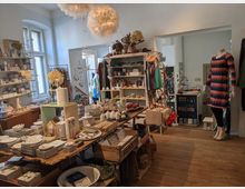 Ein kleiner Laden mit Holzboden, gefüllt mit Regalen und Tischen, die diverse Dekorationsartikel, Keramik, Kleidung und Geschenke präsentieren. Links sind große Fenster, die Tageslicht einlassen, und im Raum hängen dekorative Federlampen von der Decke.