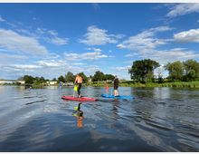 Mehrere Personen stehen auf Paddleboards auf einem ruhigen Fluss, umgeben von grüner Uferlandschaft mit Bäumen und einem blauen Himmel mit einigen Wolken. Im Hintergrund sind eine Brücke und vereinzelte Gebäude zu sehen.