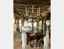 Ein rustikaler Veranstaltungsraum mit Holzgebälk, dekoriert mit Lichterketten an der Decke. Im Vordergrund ein gedeckter Tisch mit einem silbernen Kerzenleuchter, Blumenarrangements und gefalteten weißen Stoffservietten.