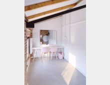 Minimalistischer, heller Raum mit schräger Holzdecke und Sichtbalken, einer Backsteinwand links und weißen Wänden. Ein schlichter weißer Tisch steht an der Wand, davor zwei rosa Stühle, ergänzt durch einen runden Spiegel und einen kleinen rosafarbenen Mülleimer.