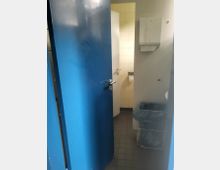 Tür zu einer öffentlichen Toilette mit blauer Außenseite, die einen teilweise sichtbaren Waschraum mit gelblich gefliesten Wänden zeigt. Rechts im Vordergrund befinden sich ein Papierhandtuchspender und ein Abfalleimer mit Drahtgestell.