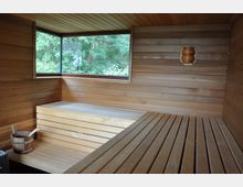 Innenraum einer modernen Sauna aus hellem Holz mit horizontalen Holzvertäfelungen. Ein großes Panoramafenster bietet Blick auf grüne Bäume, daneben ein Saunaeimer mit Kelle auf der Sitzbank.