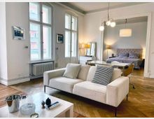 Wohnbereich einer modernen Wohnung mit Holzboden, hellen Wänden und großen Fenstern. Im Vordergrund ein cremefarbenes Sofa, ein Couchtisch mit Dekorationen, und im Hintergrund ein Schlafzimmer mit einem Bett, Nachttischen und Lampen.