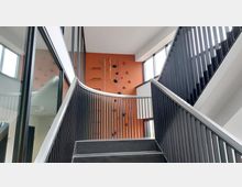 Treppenhaus mit schwarzen Geländern und großzügigen Fenstern, dahinter eine orangefarbene Wand mit Klettergriffen und Kletterseilen.