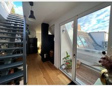Moderne Dachgeschosswohnung mit einer Holztreppe auf der linken Seite, großen Glastüren auf der rechten Seite, die zu einem Balkon mit Pflanzen und Blick auf benachbarte Dachfenster führen. Der Innenraum zeigt Holzboden, schwarze Möbelelemente und eine helle, minimalistische Einrichtung.