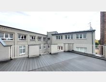 Flachdach-Terrasse mit grauem Bodenbelag und weißer Metall-Brüstung, umgeben von grau verputzten Gebäudefassaden mit rechteckigen Fenstern. Im Hintergrund ist ein gemauerter Schornstein sowie ein minimal bewölkter Himmel sichtbar.