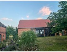 Backsteingebäude mit rotem Ziegeldach in einer ländlichen Umgebung, umgeben von Bäumen und einem Garten mit hohem Gras im Vordergrund. Rechts und links des Gebäudes stehen weitere Backsteinhäuser, während der Himmel klar ist mit einigen rosa Wolken.