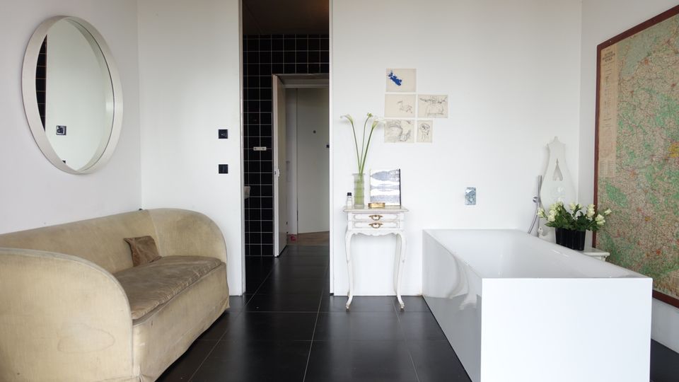 Minimalistisch eingerichteter Raum mit schwarzem Fliesenboden, einem beigen Sofa, einer freistehenden weißen Badewanne und einem kleinen weißen Tisch. An der weißen Wand hängen Zeichnungen und ein Rundspiegel, während auf der rechten Seite eine große Landkarte sichtbar ist.