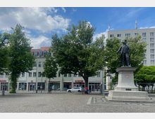 Statue auf einem gepflasterten Platz mit Bäumen im Vordergrund, flankiert von mehrstöckigen Gebäuden mit Geschäften im Erdgeschoss. Der Himmel ist teils bewölkt, und eine Person zieht einen Wagen auf der rechten Bildseite.