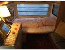 Innenansicht eines Wohnwagens mit einem schmalen Bett an der Rückwand, umgeben von Fenstern. Im Vordergrund steht ein kleiner Schrank mit einer Lampe, einem Spiegel, Teelichtern und einer Pflanze.