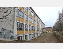 Langer, mehrstöckiger Gebäuderiegel mit Rasterfassade aus vielen Fenstern und gelb-weißen Fassadenelementen, an der Stirnseite mit Graffiti. Davor liegt ein schmaler Grünstreifen mit Weg und kahlen Bäumen; im Hintergrund ist ein weiteres Backsteingebäude zu sehen.