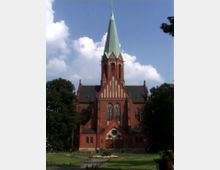 Backsteinkirche mit hohem grünen Turm und gotischen Elementen, umgeben von Bäumen und einer gepflegten Wiese mit Blumenbeeten im Vordergrund. Der Himmel ist klar mit wenigen Wolken.