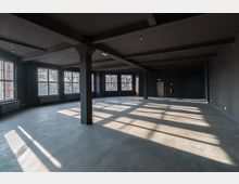 Ein leerer, großer Raum mit schwarzer Decke und Wänden, grauem Betonboden und mehreren großzügigen Fenstern, die Tageslicht hereinlassen. Massive schwarze Säulen und sichtbare Deckenträger dominieren die industrielle Architektur.