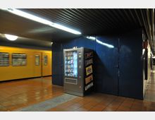 Untergrundbahnhof mit gelbem U-Bahn-Wagen im Hintergrund, einem Snackautomaten an einer blauen Wand und einem Boden aus orangefarbenen Fliesen. потолок mit beleuchteten Metallpaneelen.