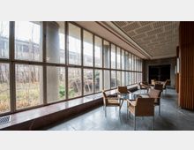 Ein Innenraum mit großen, bodentiefen Fenstern, die einen Blick auf einen verwilderten Außenbereich mit Sträuchern und Bäumen bieten. Der Raum ist mit marmorgefliesten Böden, schlichten Holzwänden und Sitzgruppen aus geflochtenen Stühlen und kleinen Tischen eingerichtet.