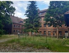 Backsteingebäude mit verwitterten Fenstern, umgeben von hohen Bäumen und einer Wiese mit ungemähtem Gras. Im Vordergrund ein kleiner Zaun und ein mit Kopfsteinpflaster bedeckter Weg.