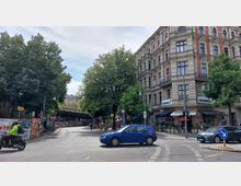 Straßenszene mit einem Eckgebäude mit verzierten Fassaden und Geschäften im Erdgeschoss, darunter ein Laden mit der Aufschrift "OVERKILL". Links des Gebäudes steht eine Reihe großer Bäume, und im Hintergrund ist eine Hochbahnbrücke mit einer gelben U-Bahn zu sehen.