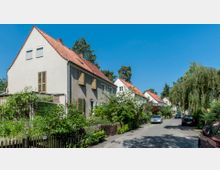 Eine ruhige Wohnstraße mit Reihenhäusern im traditionellen Stil, gekennzeichnet durch Satteldächer mit roten Ziegeln und hell verputzte Fassaden. Vorgärten mit üppiger Bepflanzung und ein paar geparkte Autos säumen die Straße, umgeben von Bäumen und Grünflächen.