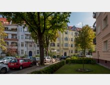 Straßenszene mit mehrstöckigen Wohnhäusern im Altbaustil, darunter ein gelbes Gebäude mit Rundbogenfenstern. Im Vordergrund stehen geparkte Autos unter Bäumen, daneben eine kleine Grünfläche mit Rasen und niedriger Hecke.