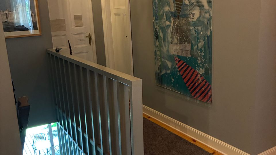 Schmaler Treppenhausflur in einem Wohnhaus mit grauen Wänden, Holzboden und einem Geländer zur Treppe nach unten. An den Wänden hängen gerahmte Bilder, darunter ein großes vertikales Kunstwerk, und neben zwei weißen Innentüren ist eine Wandlampe angebracht.