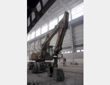 Ein gelber Bagger mit großen Reifen steht in einer geräumigen Industriehalle mit hohen Decken und großen Oberlichtern. Im Hintergrund ist eine Person neben Baustellenequipment zu sehen.