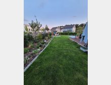 Großer, gepflegter Hausgarten mit breitem Rasenstreifen, links mit Beeten und jungen Bäumen bepflanzt und rechts mit Holzterrasse sowie Sonnenschirm. Im Hintergrund stehen moderne Wohnhäuser, und in der Mitte des Gartens befindet sich ein Holzgestell mit Hängematte.