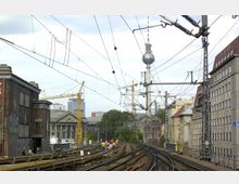 Blick auf eine erhöhte Bahntrasse in einer städtischen Umgebung mit umliegenden Wohn- und Industriegebäuden, Baukränen im Hintergrund und dem Berliner Fernsehturm im Zentrum. Arbeiter in orangefarbenen Westen führen Bau- oder Wartungsarbeiten auf den Schienen durch.