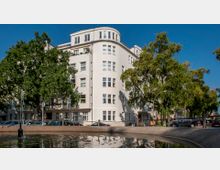 Wohnhaus in einem urbanen Umfeld mit weißer Fassade, großen Fenstern und abgerundeter Ecke. Im Vordergrund ein kleiner Teich mit sprudelndem Brunnen, umgeben von Bäumen und geparkten Autos.