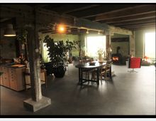 Offener Wohnraum in einem industriellen Stil mit sichtbaren Holzbalken und Betonboden. Die Einrichtung umfasst eine Küchenzeile, einen Esstisch mit Stühlen, große Zimmerpflanzen, einen gemütlichen Holzofen und große Fenster, die Tageslicht einlassen.