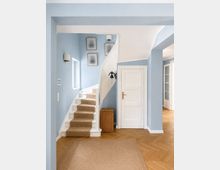 Heller Eingangsbereich in einem Wohnhaus mit hellblauen Wänden, weißer Tür und geschwungener Treppe mit Teppichläufer. Holzparkett in Fischgrätmuster, kleiner Teppich, Wandlampen, gerahmte Bilder und ein Korb neben der Treppe.