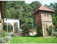 Ein gepflegter Garten mit einem Backsteinturm mit roten Dachziegeln und einer Holztür in der Vordergrundmitte. Links befindet sich ein angrenzender Wintergarten mit Glasfront und Holzrahmen, umgeben von Blumen, Pflanzen und grüner Rasenfläche, mit Bäumen im Hintergrund.