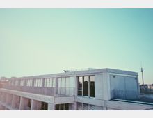 Ein modernes Gebäude mit flacher Fassade aus Beton, großen Fenstern und Balkongittern, im Hintergrund ist der Berliner Fernsehturm vor klarem, bläulichem Himmel sichtbar.