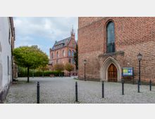 Historischer Platz mit Pflastersteinen, umgeben von Backsteingebäuden. Im Vordergrund ein gotischer Eingangsbereich einer Kirche, darüber ein großes, spitzbogiges Fenster; im Hintergrund ein weiteres rotes Backsteingebäude mit verzierten Giebeln und mehreren Bäumen.