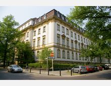 Ein großes, dreistöckiges historisches Gebäude mit heller Fassade, gelblichen Akzenten und verzierten Fensterelementen, umgeben von Bäumen und parkenden Autos. Im Vordergrund befindet sich eine Straßenszene mit Verkehrszeichen und Gehweg.