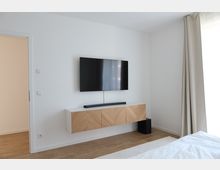 Minimalistisch eingerichtetes Zimmer mit einem an der Wand montierten Flachbildfernseher, einem hängenden Holzschrank mit Fischgrätenmuster und einem Lautsprecher darunter. Rechts hängt ein beiger Vorhang über einem bodentiefen Fenster, und der Boden ist aus Holz.